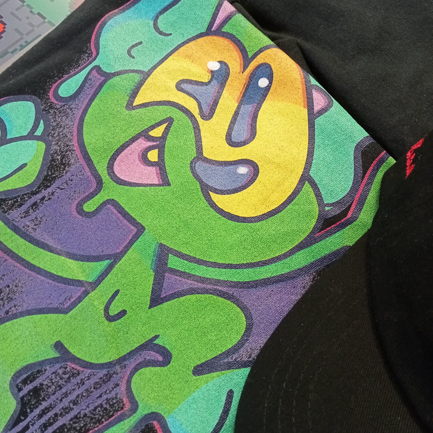 Green slime graffiti character by streetwear brand CORKiE, printed onto a black shirt with a spray paint effect in the background.